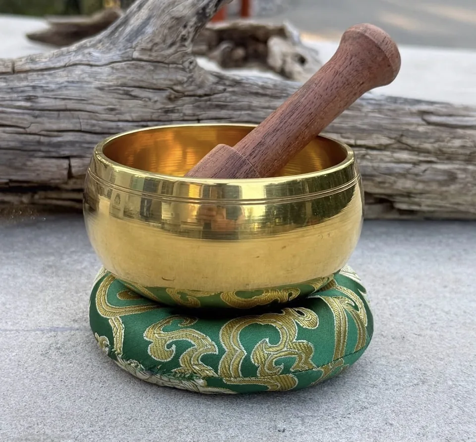 A polished singing bowl resting on a green brocade cushion with its wooden mallet
