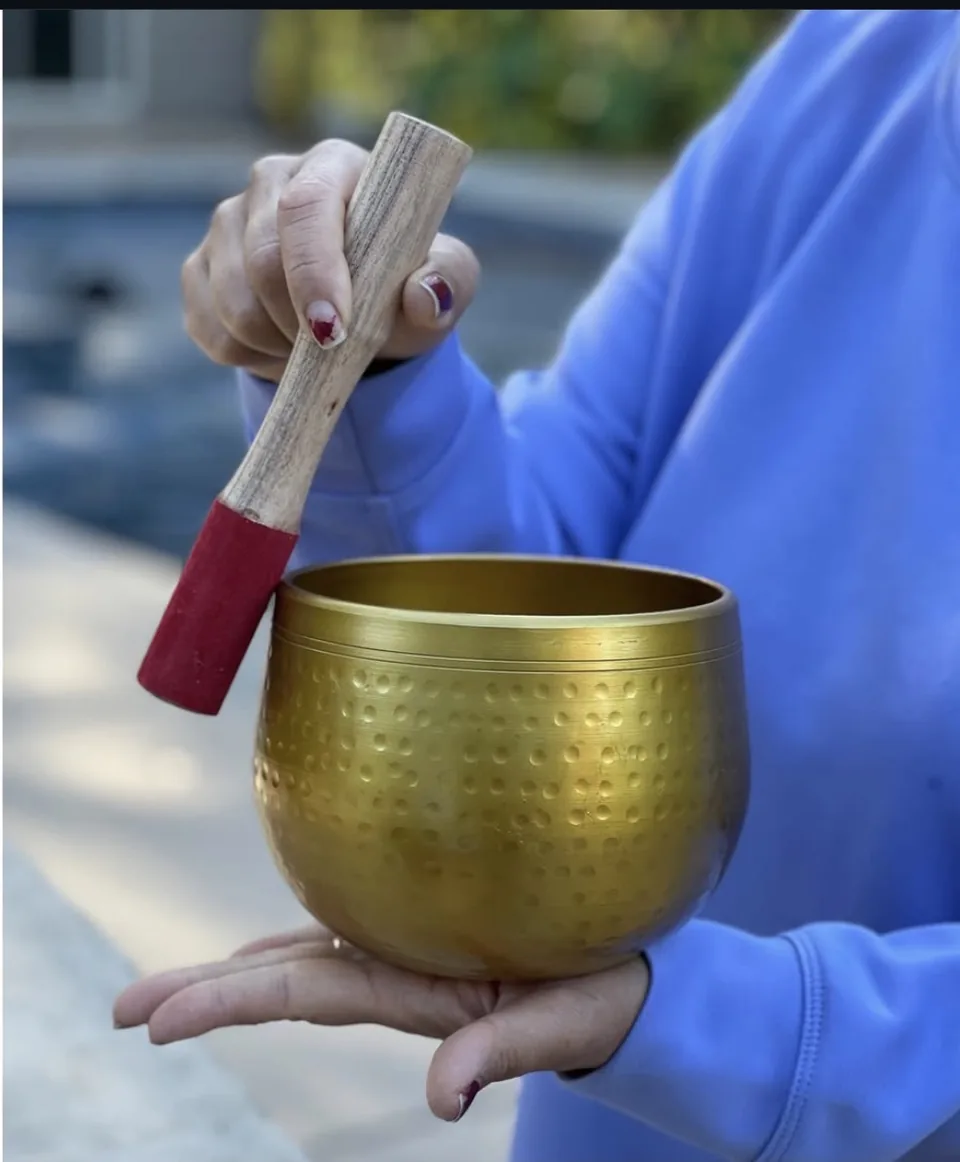 Hands striking a hammered brass singing bowl with a red-tipped mallet